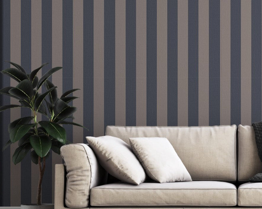 A modern living room features a dark navy blue and muted grey-brown striped wallpaper.
The design showcases clean, evenly spaced vertical stripes filling the entire back wall.
In front, a beige fabric sofa with decorative pillows rests on a chevron pattern rug.
The contemporary, stylish space is finished with a large potted green plant on the left.
