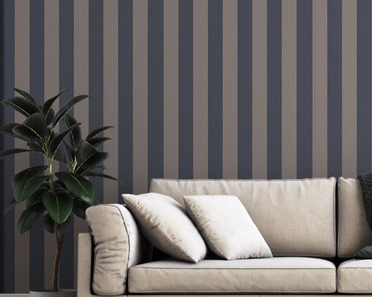 A modern living room features a dark navy blue and muted grey-brown striped wallpaper.
The design showcases clean, evenly spaced vertical stripes filling the entire back wall.
In front, a beige fabric sofa with decorative pillows rests on a chevron pattern rug.
The contemporary, stylish space is finished with a large potted green plant on the left.