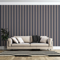 A modern living room features a dark navy blue and muted grey-brown striped wallpaper.
The design showcases clean, evenly spaced vertical stripes filling the entire back wall.
In front, a beige fabric sofa with decorative pillows rests on a chevron pattern rug.
The contemporary, stylish space is finished with a large potted green plant on the left.