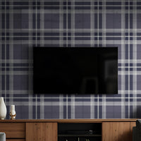 A modern living room features a classic plaid wallpaper in deep charcoal and light grey tones.
The detailed fabric-look design showcases an intricate grid pattern of intersecting lines across the wall.
In front, a unique dark grey tufted accent chair with angled wooden legs is positioned to the right.
The stylish setup is completed by a round white side table holding minimalist vases and decorative items.