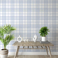 A bright interior space features a soft ivory and cornflower blue plaid wallpaper.
The detailed design shows thin blue lines intersecting to form clean grids across a white base.
In front, a rustic wooden console table holds modern ceramic vases and candles.
The breezy, minimalist setup is accented with vibrant green potted plants on both sides.