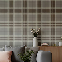 A modern entryway hallway features a classic truffle tartan plaid wallpaper in rich chocolate brown and cream tones.
The textured fabric-look design displays a bold grid pattern with intersecting brown and off-white bands.
In front, a minimalist light wooden console table stands next to a tall, dark wood interior door.
The warm, inviting space is completed by a round white globe lamp and a small decorative stool.