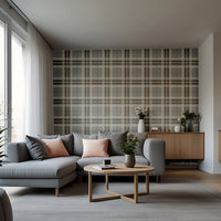 A modern entryway hallway features a classic truffle tartan plaid wallpaper in rich chocolate brown and cream tones.
The textured fabric-look design displays a bold grid pattern with intersecting brown and off-white bands.
In front, a minimalist light wooden console table stands next to a tall, dark wood interior door.
The warm, inviting space is completed by a round white globe lamp and a small decorative stool.