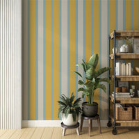 A lively room setup features a vintage oasis yellow and blue striped wallpaper.
The detailed design shows alternating vertical bands of golden yellow, muted teal, and beige.
In front, leafy green potted plants sit neatly on small wooden plant stands.
To the right, a rustic wooden bookshelf on wheels holds books and decorative items.