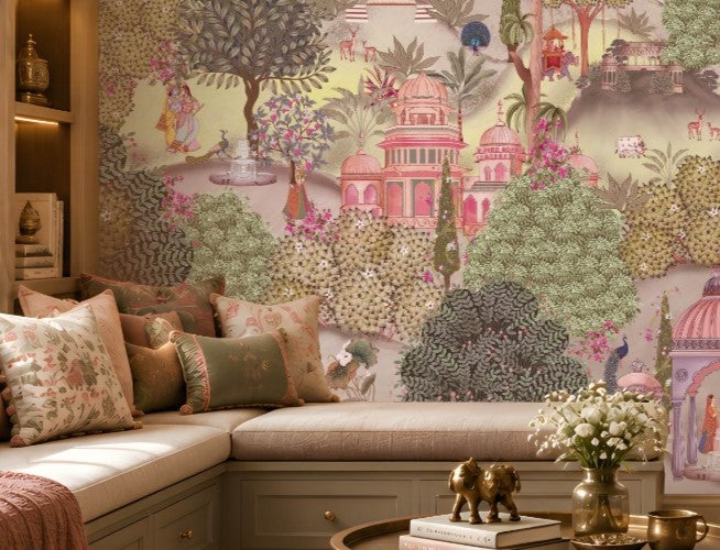 A cozy seating nook with a cushioned bench decorated with patterned pillows in beige, pink, and green. In front sits a small table holding two golden elephant figurines and a vase of white flowers. Behind, a large mural depicts a lush garden with domed pavilions, arches, flowering trees, and figures in traditional attire, creating an elegant and culturally inspired atmosphere.
