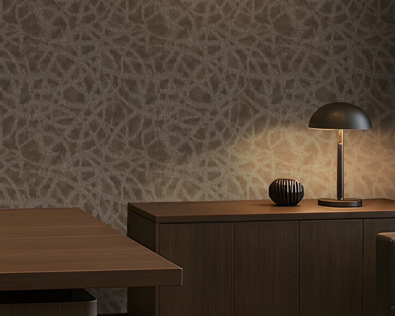 A sophisticated executive office with a warm, contemporary aesthetic: a large walnut desk, dark leather seating, integrated wood cabinetry, a textured taupe-gray feature wall, concealed warm LED lighting, recessed ceiling illumination, floor-to-ceiling windows with roller shades, and a refined lounge area with a leather sofa and marble coffee table. The palette is restrained and architectural, combining dark wood, charcoal, warm gray, and brass accents.
