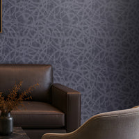 "Modern living room featuring a premium slate blue seamless texture wallpaper with an elegant organic abstract pattern that adds depth and visual interest to the walls. The sophisticated interior includes a dark brown leather sofa, two upholstered accent chairs, a black marble coffee table, warm brass table lamp, built-in wooden shelving with ambient lighting, abstract wall art, and floor-to-ceiling curtains. This luxurious textured wallpaper is ideal for contemporary