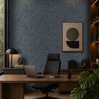 A modern, moody home office featuring a dark blue textured accent wall, warm walnut cabinetry and desk, black leather office chair, minimalist abstract artwork, integrated shelving with warm LED lighting, and abundant greenery. Natural light enters from the tall window on the left, complemented by warm recessed ceiling lights. The overall aesthetic is sophisticated, contemporary, and calm, with earthy materials and muted tones.
