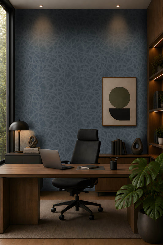A modern, moody home office featuring a dark blue textured accent wall, warm walnut cabinetry and desk, black leather office chair, minimalist abstract artwork, integrated shelving with warm LED lighting, and abundant greenery. Natural light enters from the tall window on the left, complemented by warm recessed ceiling lights. The overall aesthetic is sophisticated, contemporary, and calm, with earthy materials and muted tones.
