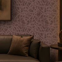 A luxurious contemporary lounge with a symmetrical layout, featuring a dark leather sofa and two matching armchairs around a low rounded rectangular marble coffee table. The room has a warm brown and taupe palette, dark wood built-in shelving, patterned textured wallpaper, brass-accented lighting, sculptural décor, and a large abstract artwork centered on the feature wall. Concealed cove lighting and warm wall sconces create an intimate, sophisticated atmosphere.