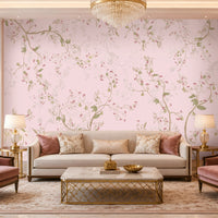 Blush Pink floral wallpaper featuring an elegant botanical design with delicate soft pink blossoms, muted green leaves, graceful winding branches, and subtle ivory floral detailing, creating a sophisticated and seamless visual flow across the feature wall. The wallpaper is showcased in a luxurious contemporary living room with a soft neutral sofa, blush pink velvet accent chairs, decorative cushions, gold-finished side tables, elegant table lamps, a statement crystal chandelier.