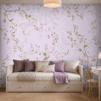 "A serene bedroom with a white daybed topped with purple and cream pillows and a lavender throw blanket. A floral pastel wallpaper covers the wall behind, featuring delicate vines in lavender, green, and pink. To the right, a white desk with a wooden chair, lamp, and vase of flowers sits near a window with Roman shades. Built-in shelves on the left display books and decorative items. Soft natural light and a ceiling lamp create a calm, elegant atmosphere."