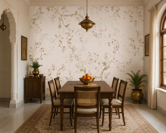 Blossom Veil Mural – Dusty Rose Pink floral wallpaper displayed across an elegant Indian-inspired dining room, featuring delicate pastel pink and peach blossoms intertwined with warm ivory vines, soft botanical branches, muted green leaves, and fine floral detailing on a subtle dusty rose pink background. The graceful floral composition flows seamlessly across the feature wall, creating a refined and sophisticated look. The room includes a traditional wooden dining table with woven 