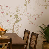 Blossom Veil Mural – Dusty Rose Pink floral wallpaper displayed across an elegant Indian-inspired dining room, featuring delicate pastel pink and peach blossoms intertwined with warm ivory vines, soft botanical branches, muted green leaves, and fine floral detailing on a subtle dusty rose pink background. The graceful floral composition flows seamlessly across the feature wall, creating a refined and sophisticated look. The room includes a traditional wooden dining table with woven 