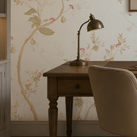 "A cozy home office with a wooden desk, beige swivel chair, brass desk lamp, and books neatly arranged. A framed landscape artwork hangs above the desk against light floral wallpaper. Built-in shelves with books and decor sit to the left, while a window on the right brings in natural light. A neutral rug covers the floor, complementing the warm and elegant aesthetic."  