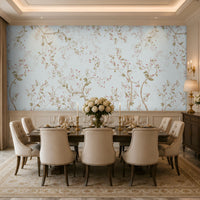 An elegant dining room featuring a long wooden table set with fine dinnerware, gold candlesticks, and a white rose centerpiece. Ten upholstered beige chairs surround the table. The walls are covered in light blue floral wallpaper, with a dark wooden sideboard and lamps on the right beneath a large mirror. Natural light filters through tall windows with beige curtains, illuminating the patterned rug and refined, classic décor.  