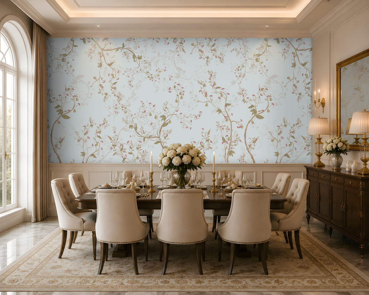 An elegant dining room featuring a long wooden table set with fine dinnerware, gold candlesticks, and a white rose centerpiece. Ten upholstered beige chairs surround the table. The walls are covered in light blue floral wallpaper, with a dark wooden sideboard and lamps on the right beneath a large mirror. Natural light filters through tall windows with beige curtains, illuminating the patterned rug and refined, classic décor.  