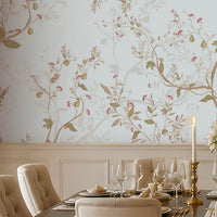 An elegant dining room featuring a long wooden table set with fine dinnerware, gold candlesticks, and a white rose centerpiece. Ten upholstered beige chairs surround the table. The walls are covered in light blue floral wallpaper, with a dark wooden sideboard and lamps on the right beneath a large mirror. Natural light filters through tall windows with beige curtains, illuminating the patterned rug and refined, classic décor.  