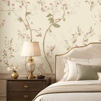 Warm ivory cream floral wallpaper with delicate botanical branches, soft pink flowers, muted sage green leaves, and subtle beige accents covering a large feature wall in an elegant luxury bedroom. The sophisticated room features a cream upholstered bed, neutral bedding, dark wooden bedside tables, classic lamps, a crystal chandelier, arched window with beige curtains, plush seating, decorative furniture, and a warm refined interior that complements the wallpaper’s soft botanical design.
