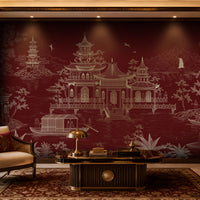 A luxury living room featuring a large wall mural with a white Chinese-style landscape and pagoda design on a deep burgundy background. The room is styled with a dark wood coffee table, a beige armchair with a decorative pillow, a patterned area rug, and warm ambient lighting.
