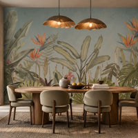 Earthy Tropical Oasis - Dusty Blue botanical wallpaper featuring lush tropical foliage, oversized bird-of-paradise flowers in warm orange, coral, and muted blue tones, and layered olive green leaves against a soft dusty blue background. Showcased in an elegant dining room with a natural wooden dining table, upholstered chairs in earthy neutral shades, warm pendant lighting, and textured décor, this tropical wallpaper brings a calming, nature-inspired charm and sophisticated elegance to contemporar