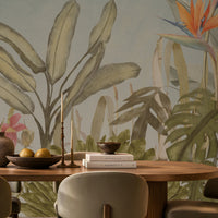 Earthy Tropical Oasis - Dusty Blue botanical wallpaper featuring lush tropical foliage, oversized bird-of-paradise flowers in warm orange, coral, and muted blue tones, and layered olive green leaves against a soft dusty blue background. Showcased in an elegant dining room with a natural wooden dining table, upholstered chairs in earthy neutral shades, warm pendant lighting, and textured décor, this tropical wallpaper brings a calming, nature-inspired charm and sophisticated elegance to contemporar