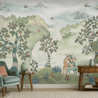 Pastel woodland wall mural featuring hand-painted sage green trees, birds in flight, and deer in a serene forest landscape, styled in a cozy living room with a green armchair and wooden console table.