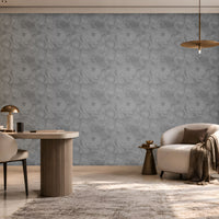 Modern, warm-toned home office or reading nook featuring a large gray patterned accent wall. On the left, built-in wooden shelving with soft LED lighting displays books, decorative objects, and a hanging plant. A rounded cream upholstered chair sits at a light-colored desk with a brass-and-marble lamp. On the right, a curved ivory lounge chair with a brown cushion and draped throw blanket is paired with a small side table. A window seat with cushions runs beneath a large window that fills the room 