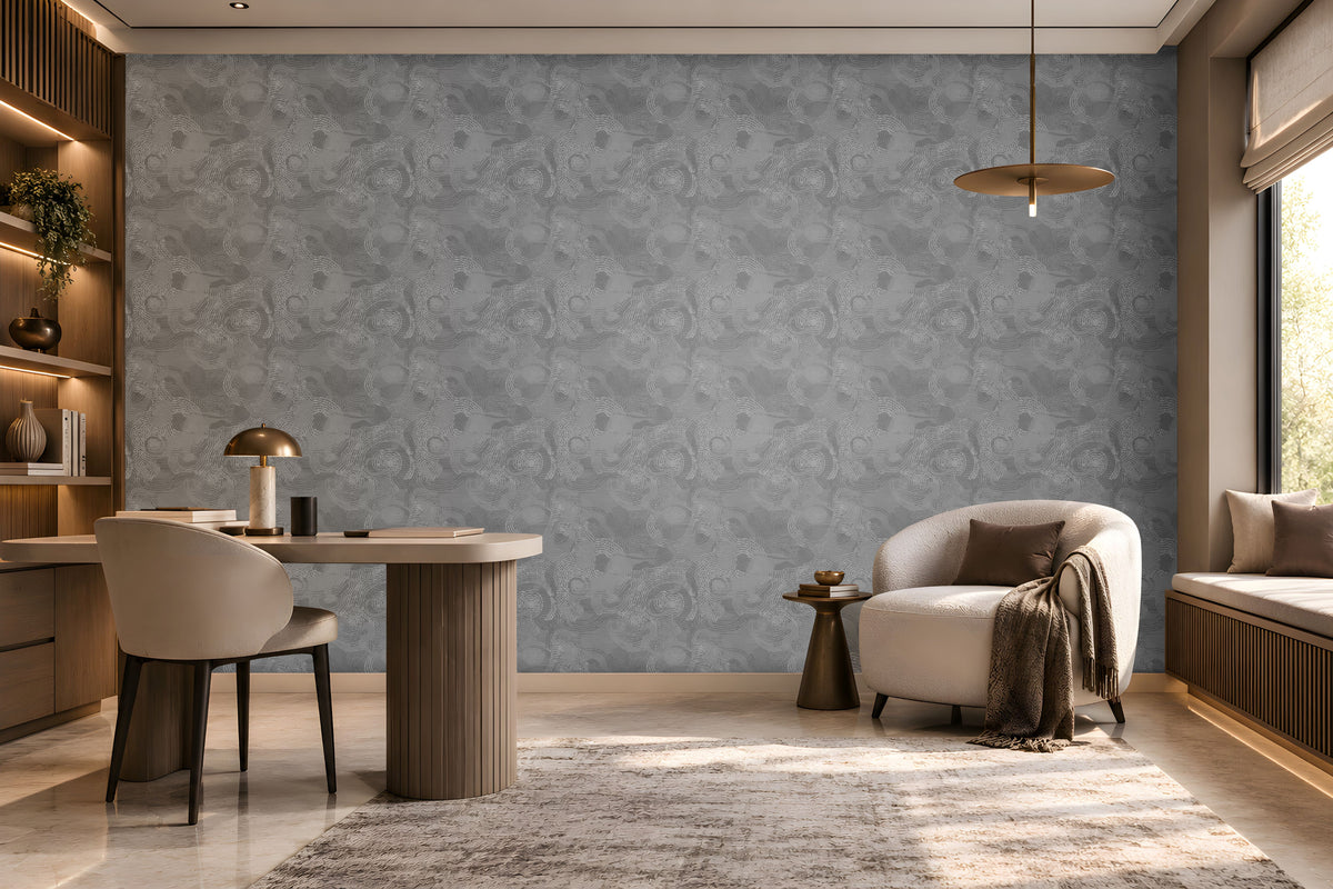 Modern, warm-toned home office or reading nook featuring a large gray patterned accent wall. On the left, built-in wooden shelving with soft LED lighting displays books, decorative objects, and a hanging plant. A rounded cream upholstered chair sits at a light-colored desk with a brass-and-marble lamp. On the right, a curved ivory lounge chair with a brown cushion and draped throw blanket is paired with a small side table. A window seat with cushions runs beneath a large window that fills the room 