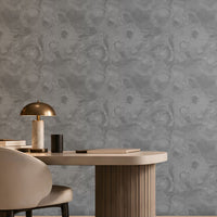 Modern, warm-toned home office or reading nook featuring a large gray patterned accent wall. On the left, built-in wooden shelving with soft LED lighting displays books, decorative objects, and a hanging plant. A rounded cream upholstered chair sits at a light-colored desk with a brass-and-marble lamp. On the right, a curved ivory lounge chair with a brown cushion and draped throw blanket is paired with a small side table. A window seat with cushions runs beneath a large window that fills the room