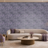 Contemporary living room with a sophisticated lavender and cream color palette. A large feature wall covered in intricate lavender-gray patterned wallpaper serves as the focal point of the space. Centered against the wall is a long, curved ivory sofa accented with purple, taupe, and textured decorative cushions. A folded lavender throw blanket drapes over the right side of the sofa. In front of the seating area, a round marble-top coffee table with a fluted bronze base displays stacked books.