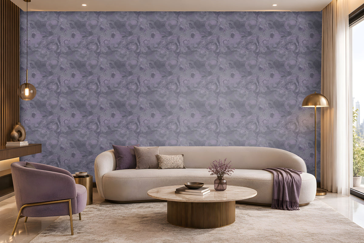 Contemporary living room with a sophisticated lavender and cream color palette. A large feature wall covered in intricate lavender-gray patterned wallpaper serves as the focal point of the space. Centered against the wall is a long, curved ivory sofa accented with purple, taupe, and textured decorative cushions. A folded lavender throw blanket drapes over the right side of the sofa. In front of the seating area, a round marble-top coffee table with a fluted bronze base displays stacked books.