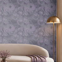 Contemporary living room with a sophisticated lavender and cream color palette. A large feature wall covered in intricate lavender-gray patterned wallpaper serves as the focal point of the space. Centered against the wall is a long, curved ivory sofa accented with purple, taupe, and textured decorative cushions. A folded lavender throw blanket drapes over the right side of the sofa. In front of the seating area, a round marble-top coffee table with a fluted bronze base displays stacked books