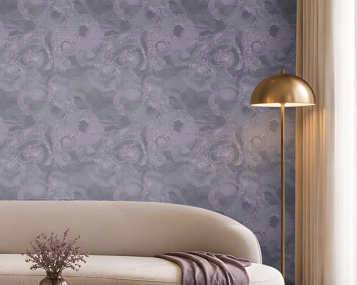 Contemporary living room with a sophisticated lavender and cream color palette. A large feature wall covered in intricate lavender-gray patterned wallpaper serves as the focal point of the space. Centered against the wall is a long, curved ivory sofa accented with purple, taupe, and textured decorative cushions. A folded lavender throw blanket drapes over the right side of the sofa. In front of the seating area, a round marble-top coffee table with a fluted bronze base displays stacked books