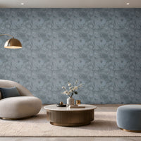 A spacious modern living room featuring a seamless blue-grey textured wallpaper with an intricate swirling pattern covering the entire accent wall. The elegant space includes a cream boucle lounge chair with a dark blue cushion, a round fluted wooden coffee table with a marble top, a matching blue-grey ottoman, and a soft beige textured rug. A sleek brass floor lamp, warm wooden display shelving with integrated lighting, and large floor-to-ceiling windows complement the wallpaper's cool tones. 
