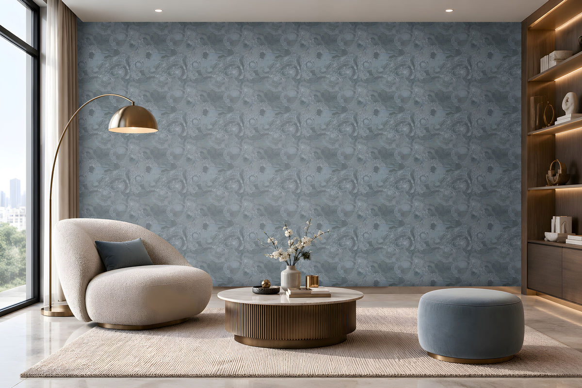 A spacious modern living room featuring a seamless blue-grey textured wallpaper with an intricate swirling pattern covering the entire accent wall. The elegant space includes a cream boucle lounge chair with a dark blue cushion, a round fluted wooden coffee table with a marble top, a matching blue-grey ottoman, and a soft beige textured rug. A sleek brass floor lamp, warm wooden display shelving with integrated lighting, and large floor-to-ceiling windows complement the wallpaper's cool tones. 