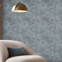 A spacious modern living room featuring a seamless blue-grey textured wallpaper with an intricate swirling pattern covering the entire accent wall. The elegant space includes a cream boucle lounge chair with a dark blue cushion, a round fluted wooden coffee table with a marble top, a matching blue-grey ottoman, and a soft beige textured rug. A sleek brass floor lamp, warm wooden display shelving with integrated lighting, and large floor-to-ceiling windows complement the wallpaper's cool tones. 