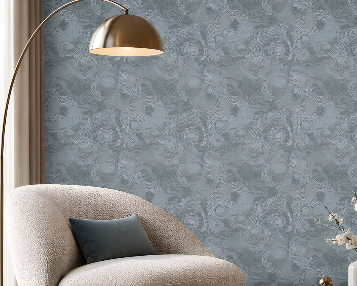A spacious modern living room featuring a seamless blue-grey textured wallpaper with an intricate swirling pattern covering the entire accent wall. The elegant space includes a cream boucle lounge chair with a dark blue cushion, a round fluted wooden coffee table with a marble top, a matching blue-grey ottoman, and a soft beige textured rug. A sleek brass floor lamp, warm wooden display shelving with integrated lighting, and large floor-to-ceiling windows complement the wallpaper's cool tones. 