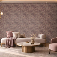 Elegant contemporary lounge featuring a mauve and dusty-rose color palette. A large accent wall covered in intricate patterned wallpaper spans the background, creating a textured focal point. In the center, a cream upholstered chaise-style sofa is decorated with blush pink, ivory, and patterned cushions, along with a draped mauve throw blanket. A round marble-top coffee table with a brushed brass base holds decorative books, a small tray, and a vase with dried stems.