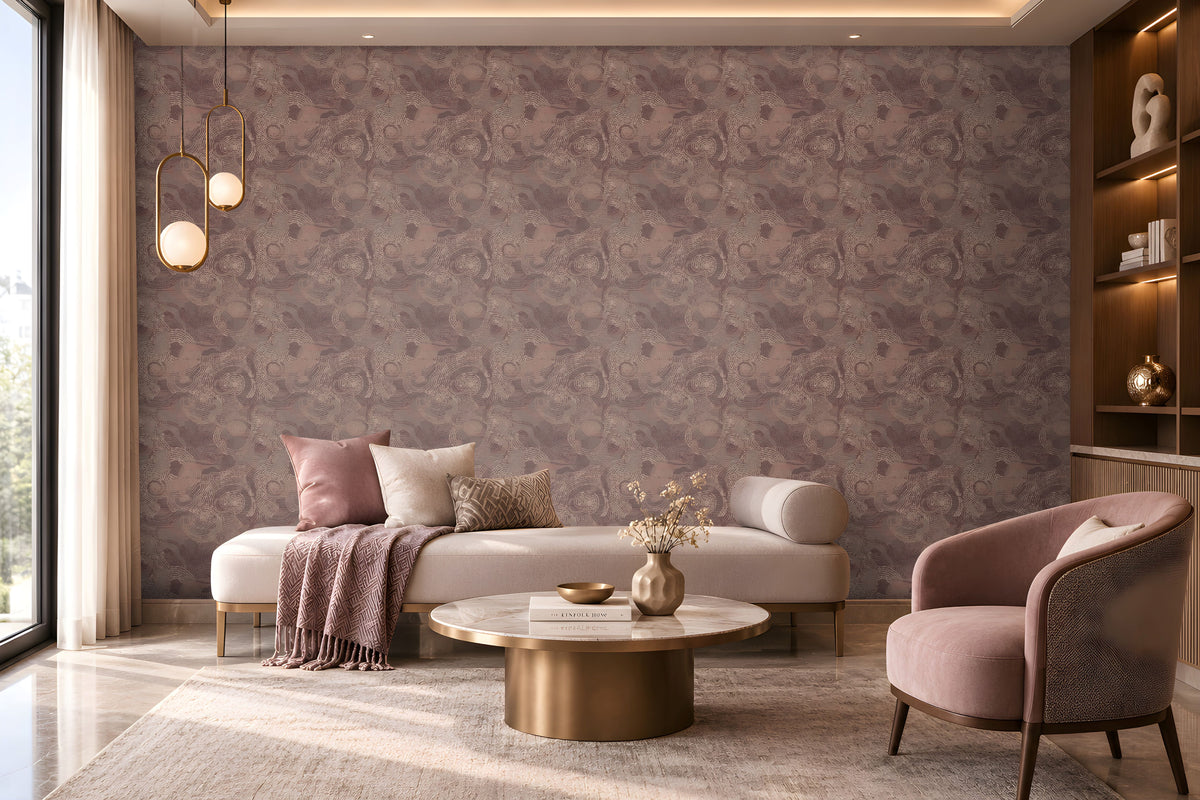 Elegant contemporary lounge featuring a mauve and dusty-rose color palette. A large accent wall covered in intricate patterned wallpaper spans the background, creating a textured focal point. In the center, a cream upholstered chaise-style sofa is decorated with blush pink, ivory, and patterned cushions, along with a draped mauve throw blanket. A round marble-top coffee table with a brushed brass base holds decorative books, a small tray, and a vase with dried stems.