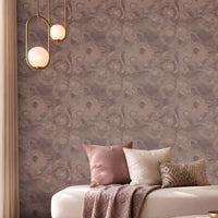 Elegant contemporary lounge featuring a mauve and dusty-rose color palette. A large accent wall covered in intricate patterned wallpaper spans the background, creating a textured focal point. In the center, a cream upholstered chaise-style sofa is decorated with blush pink, ivory, and patterned cushions, along with a draped mauve throw blanket. A round marble-top coffee table with a brushed brass base holds decorative books, a small tray, and a vase with dried stems.