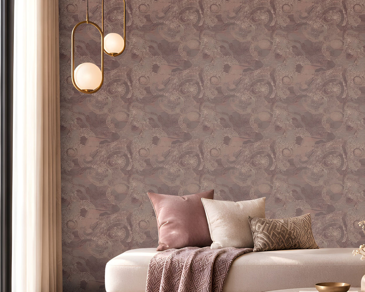 Elegant contemporary lounge featuring a mauve and dusty-rose color palette. A large accent wall covered in intricate patterned wallpaper spans the background, creating a textured focal point. In the center, a cream upholstered chaise-style sofa is decorated with blush pink, ivory, and patterned cushions, along with a draped mauve throw blanket. A round marble-top coffee table with a brushed brass base holds decorative books, a small tray, and a vase with dried stems.
