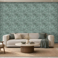 Modern minimalist living room featuring a calming teal and natural wood color palette. A large statement wall covered in textured teal wallpaper with an intricate swirling pattern forms the backdrop to the space. Positioned in front of the wall is a cream-colored sofa accented with soft green and ivory cushions, with a matching teal throw blanket draped over the right armrest.
