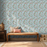 A cozy interior featuring a light blue floral wallpaper with delicate pink blossoms and green vines. A wooden woven bench with cream and blush pink cushions sits against the wall, flanked by a dark wooden side table holding a decorative metal lantern on the left and stacked cream-colored storage boxes on the right. Warm sunlight streams through sheer curtains, creating soft shadows across the wall and natural fiber rug, giving the room a calm, elegant atmosphere.
