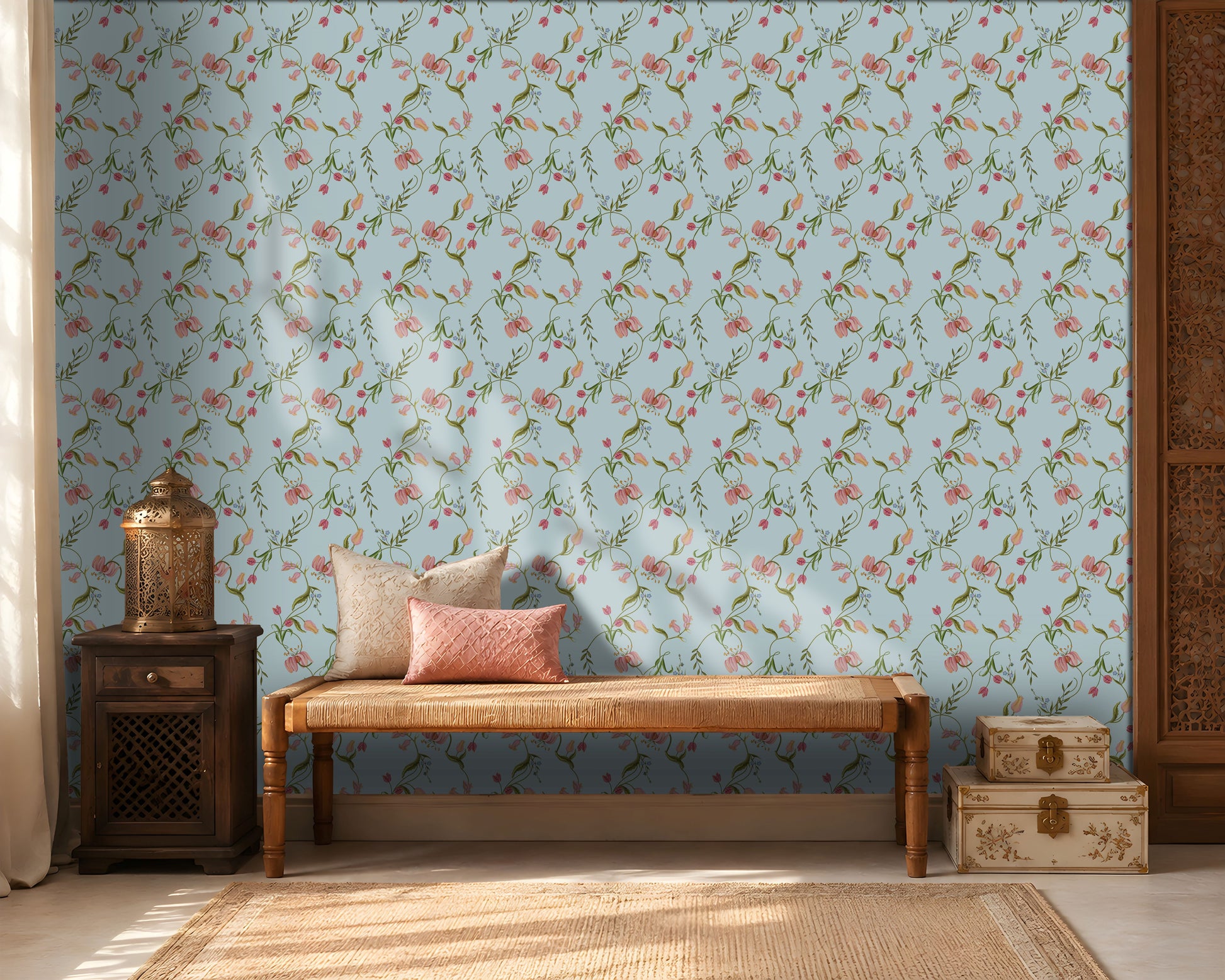 A cozy interior featuring a light blue floral wallpaper with delicate pink blossoms and green vines. A wooden woven bench with cream and blush pink cushions sits against the wall, flanked by a dark wooden side table holding a decorative metal lantern on the left and stacked cream-colored storage boxes on the right. Warm sunlight streams through sheer curtains, creating soft shadows across the wall and natural fiber rug, giving the room a calm, elegant atmosphere.
