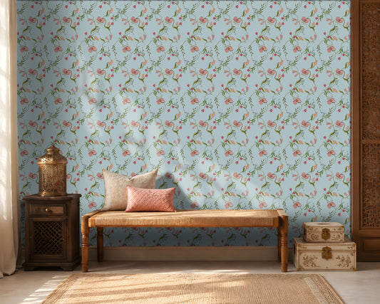 A cozy interior featuring a light blue floral wallpaper with delicate pink blossoms and green vines. A wooden woven bench with cream and blush pink cushions sits against the wall, flanked by a dark wooden side table holding a decorative metal lantern on the left and stacked cream-colored storage boxes on the right. Warm sunlight streams through sheer curtains, creating soft shadows across the wall and natural fiber rug, giving the room a calm, elegant atmosphere.
