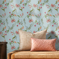 A cozy interior featuring a light blue floral wallpaper with delicate pink blossoms and green vines. A wooden woven bench with cream and blush pink cushions sits against the wall, flanked by a dark wooden side table holding a decorative metal lantern on the left and stacked cream-colored storage boxes on the right. Warm sunlight streams through sheer curtains, creating soft shadows across the wall and natural fiber rug, giving the room a calm, elegant atmosphere.
