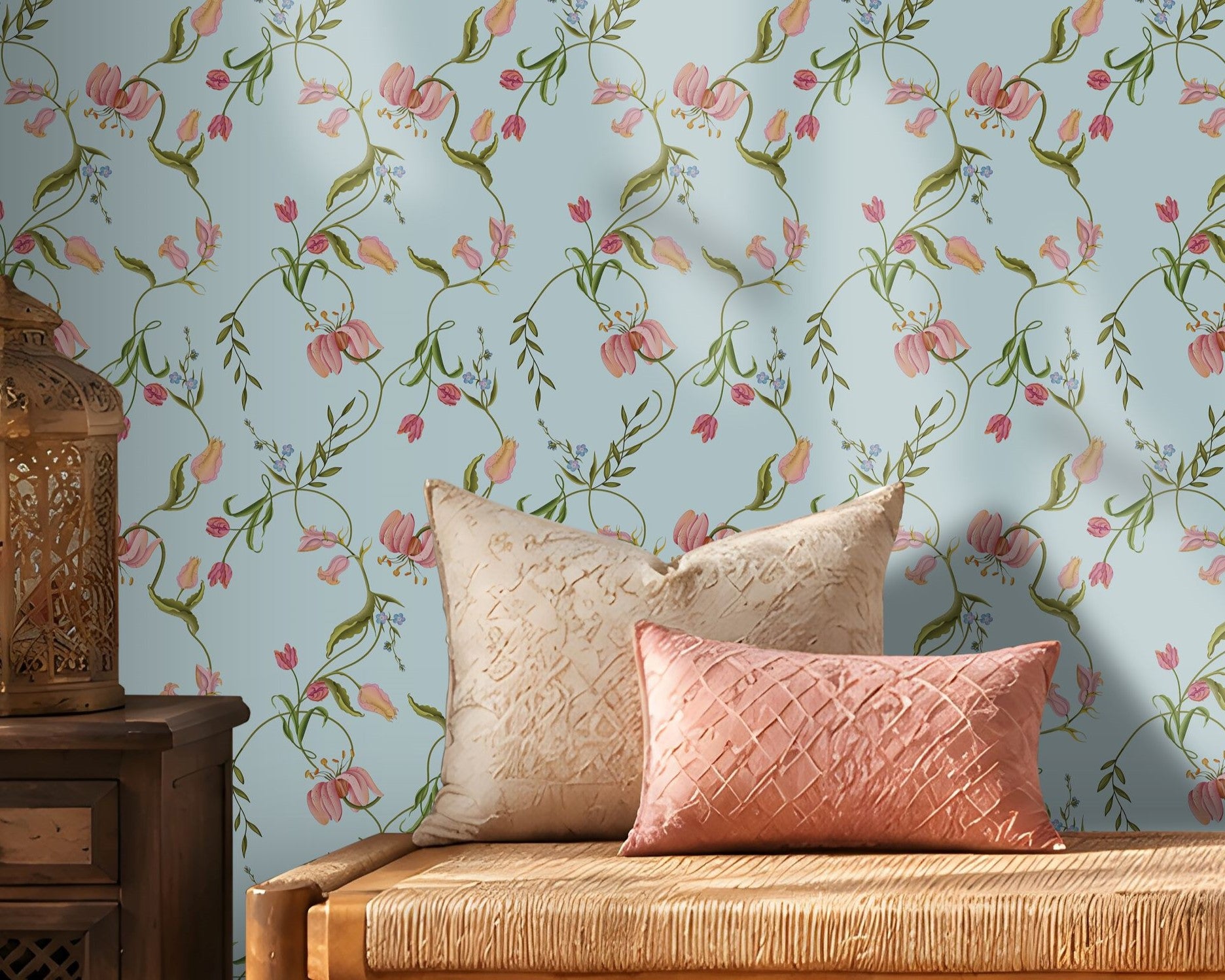 A cozy interior featuring a light blue floral wallpaper with delicate pink blossoms and green vines. A wooden woven bench with cream and blush pink cushions sits against the wall, flanked by a dark wooden side table holding a decorative metal lantern on the left and stacked cream-colored storage boxes on the right. Warm sunlight streams through sheer curtains, creating soft shadows across the wall and natural fiber rug, giving the room a calm, elegant atmosphere.