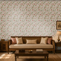 A warm and elegant living room featuring soft pink floral wallpaper with delicate green vines and blossoms. A wooden daybed-style sofa with a cane back and cream upholstery is accented with blush pink and beige cushions. Matching wooden side tables hold a decorative lantern and a table lamp, while a simple coffee table with books and a decorative bowl sits on a natural woven rug. Sunlight streams through a carved wooden window, creating soft shadows and enhancing the room's cozy, traditional ambiance.