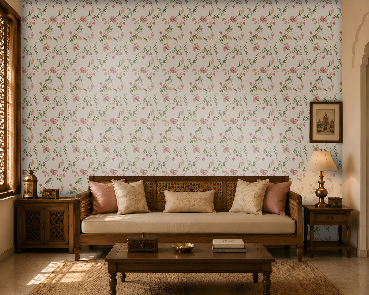 A warm and elegant living room featuring soft pink floral wallpaper with delicate green vines and blossoms. A wooden daybed-style sofa with a cane back and cream upholstery is accented with blush pink and beige cushions. Matching wooden side tables hold a decorative lantern and a table lamp, while a simple coffee table with books and a decorative bowl sits on a natural woven rug. Sunlight streams through a carved wooden window, creating soft shadows and enhancing the room's cozy, traditional ambiance.